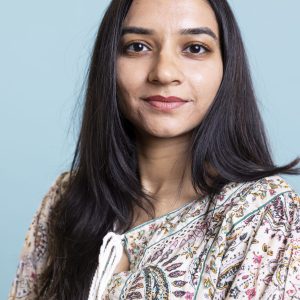 Indian woman posing in a cute stylish outfit on camera and smiling, standing against blue background. Young lively person showing confidence during a photoshoot in the studio.