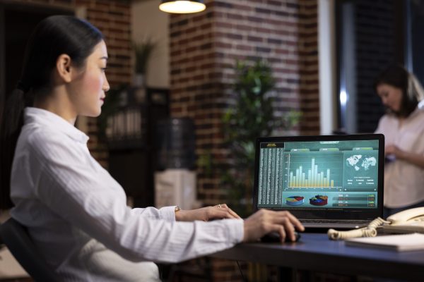 Businesswoman sitting in brick wall office, concentrated on analyzing marketing trends and company data on laptop as she works late at night. Professional analyst looking at charts displayed on screen.