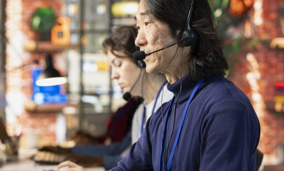 Call center agent in office helping customers by answering questions Call center agent in office helping customers by answering questions, solving their issue. Man in customer support workspace in front of PC using headset mic to talk with clients