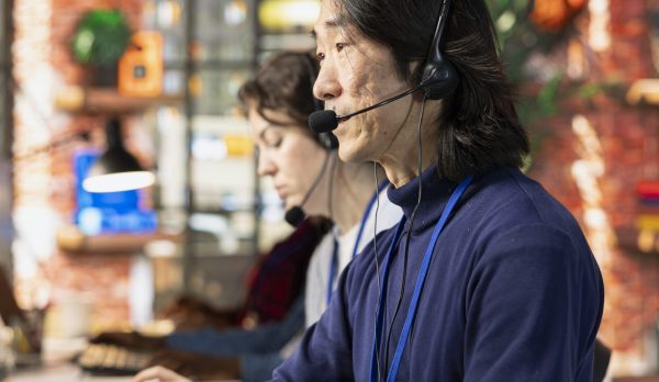 Call center agent in office helping customers by answering questions Call center agent in office helping customers by answering questions, solving their issue. Man in customer support workspace in front of PC using headset mic to talk with clients